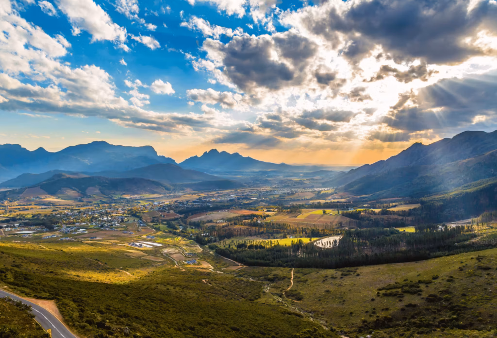 Landskaberne i Franschhoek i Sydafrika set fra oven med bjerge i baggrunden og solen, der skinner gennem mørkere skyer.
